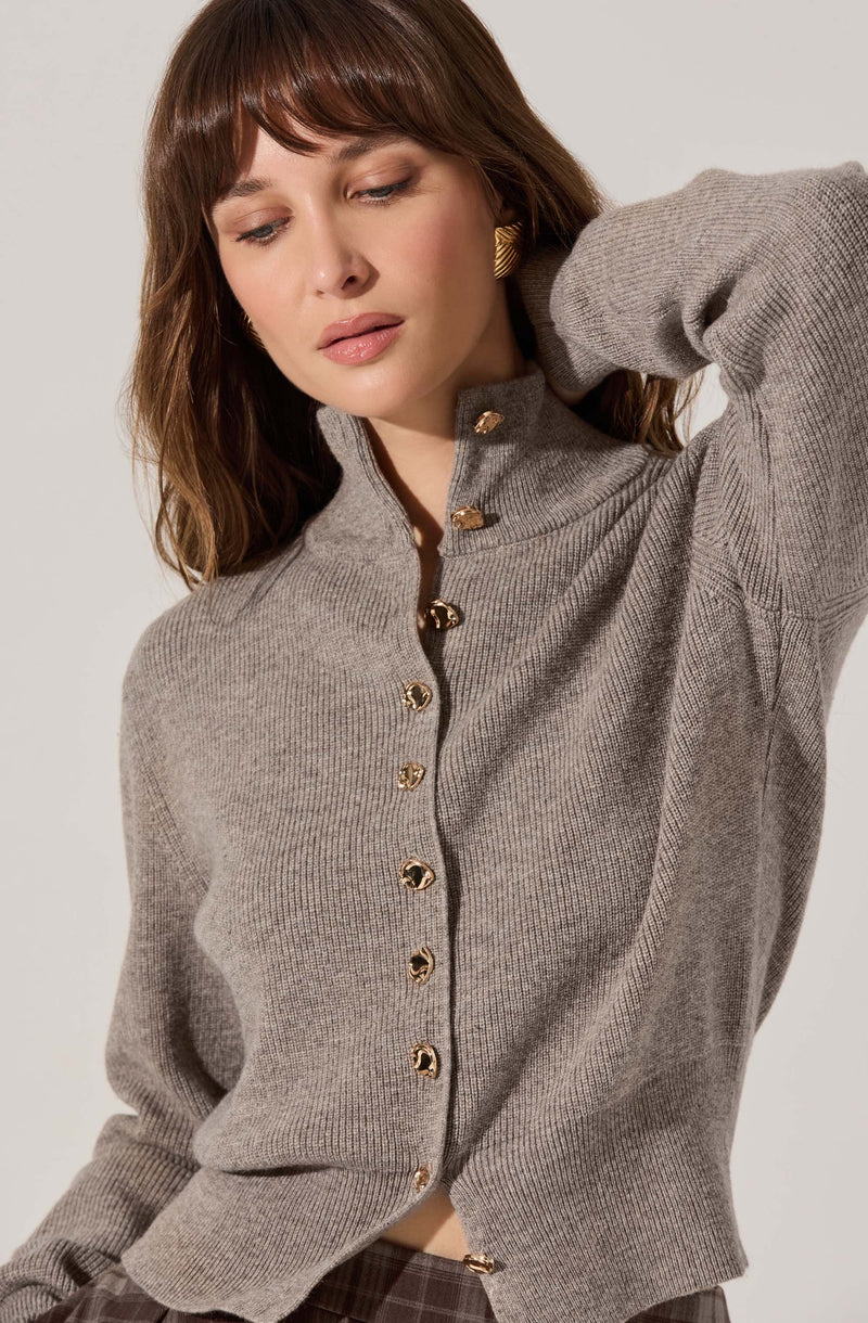 A woman is wearing a light gray, high-neck, button-up cardigan with gold buttons down the front. The cardigan has a ribbed texture at the collar, cuffs, and hem. She has chestnut brown hair with bangs and wears a gold, textured earring.