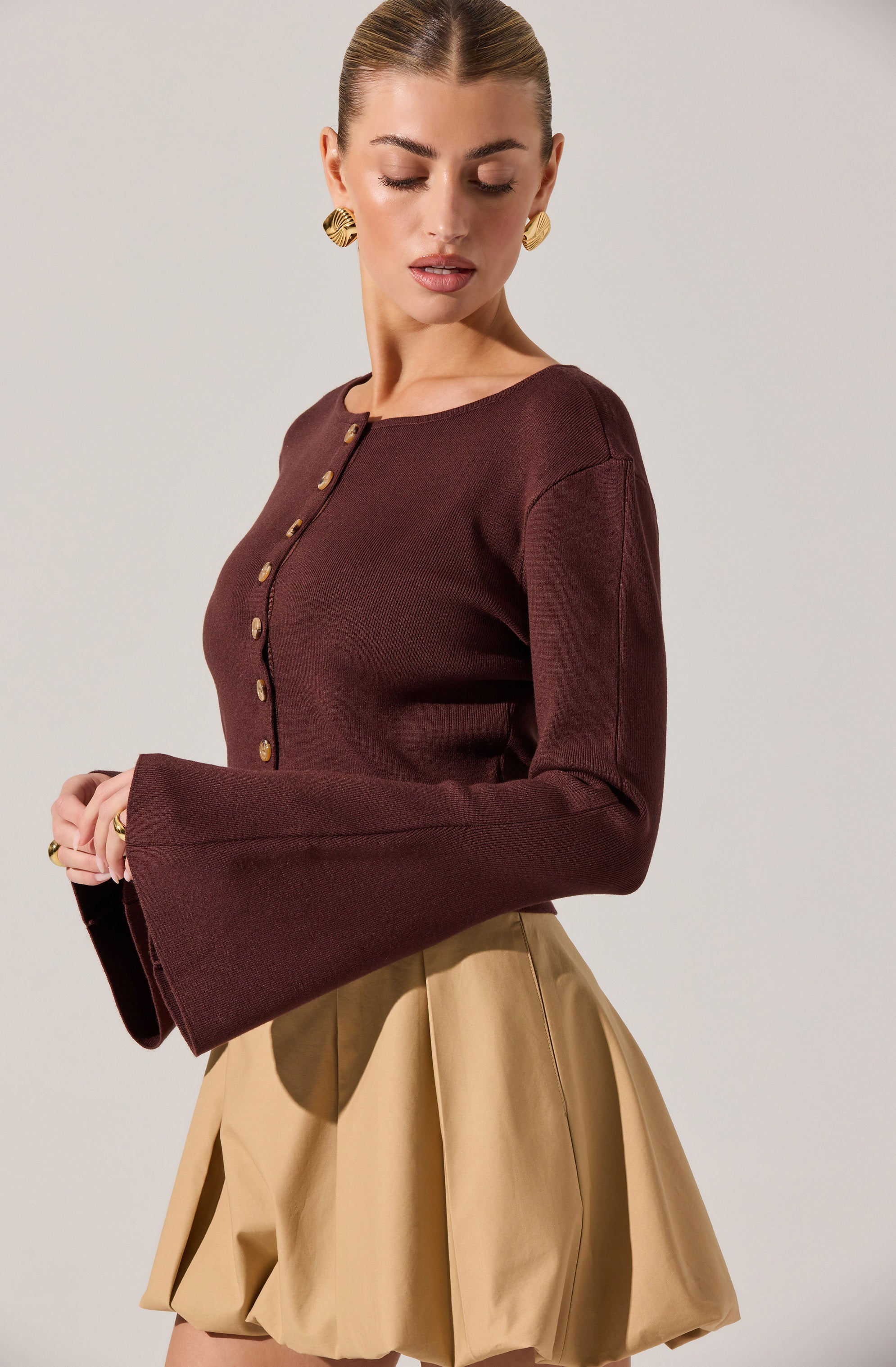 A woman is wearing a fitted, long-sleeved, burgundy top with decorative buttons running down the front. The top has wide, flared sleeves. She pairs it with high-waisted, beige, pleated shorts with a structured, voluminous look.