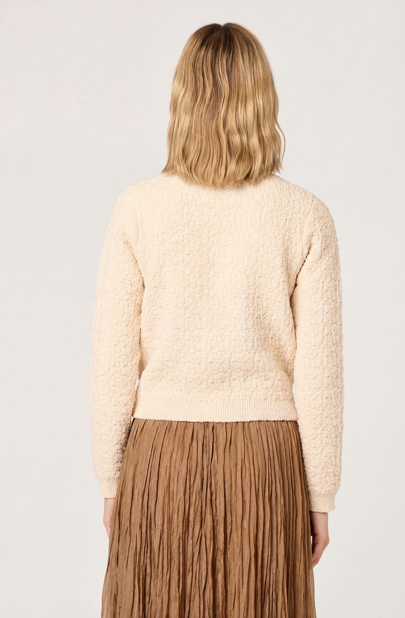 Woman wearing a cream textured sweater and a pleated brown skirt, standing with her back facing the camera.