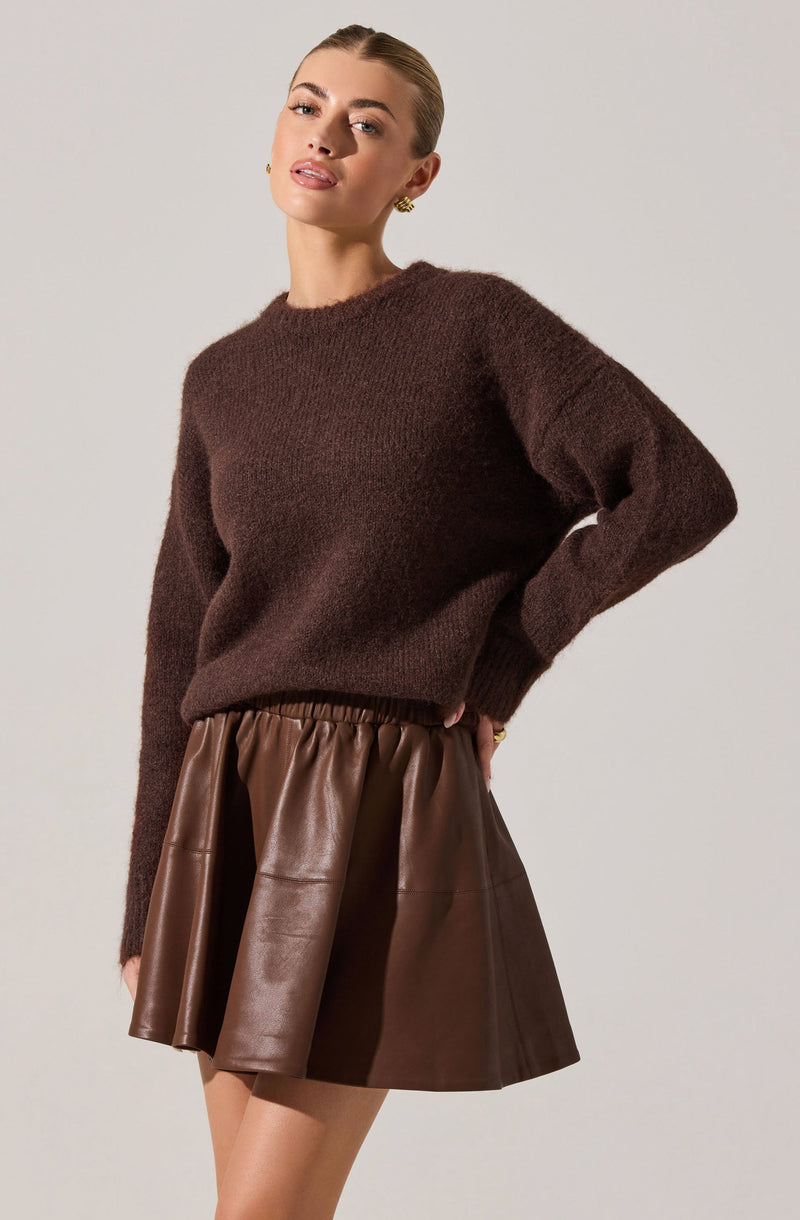 A woman is wearing a dark brown, fuzzy knit sweater with a relaxed fit and long sleeves. She pairs it with a high-waisted, brown leather skirt that has an elastic waistband and a flared, A-line silhouette.