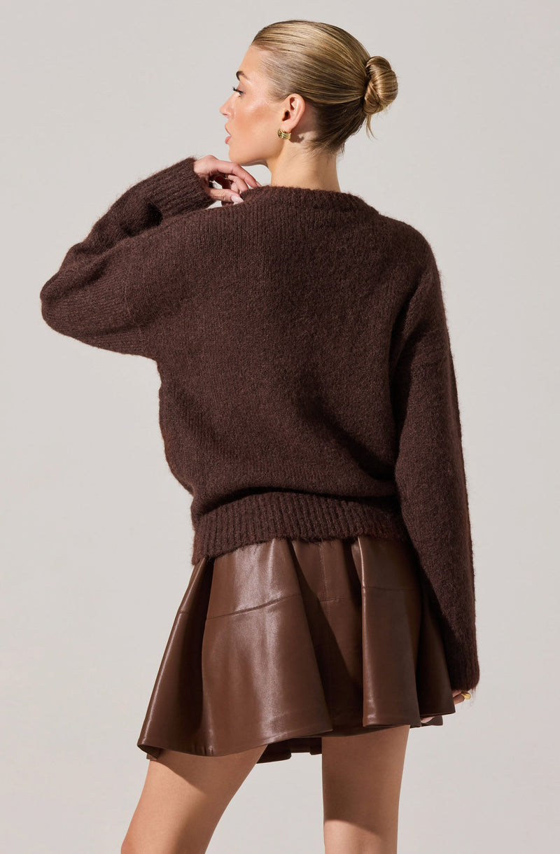 A woman is wearing a dark brown, fuzzy knit sweater with a relaxed fit and ribbed hem. She pairs it with a shiny, dark brown leather skirt that has a flared, A-line silhouette and a pleated design.