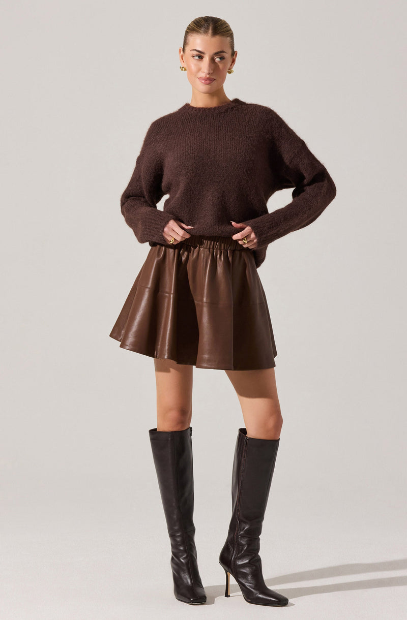 A woman is wearing a dark brown, fuzzy, long-sleeved sweater with a relaxed fit. She pairs it with a high-waisted, brown leather skirt that has a slightly flared, A-line silhouette. She completes the look with tall, black leather knee-high boots that have pointed toes and high heels.