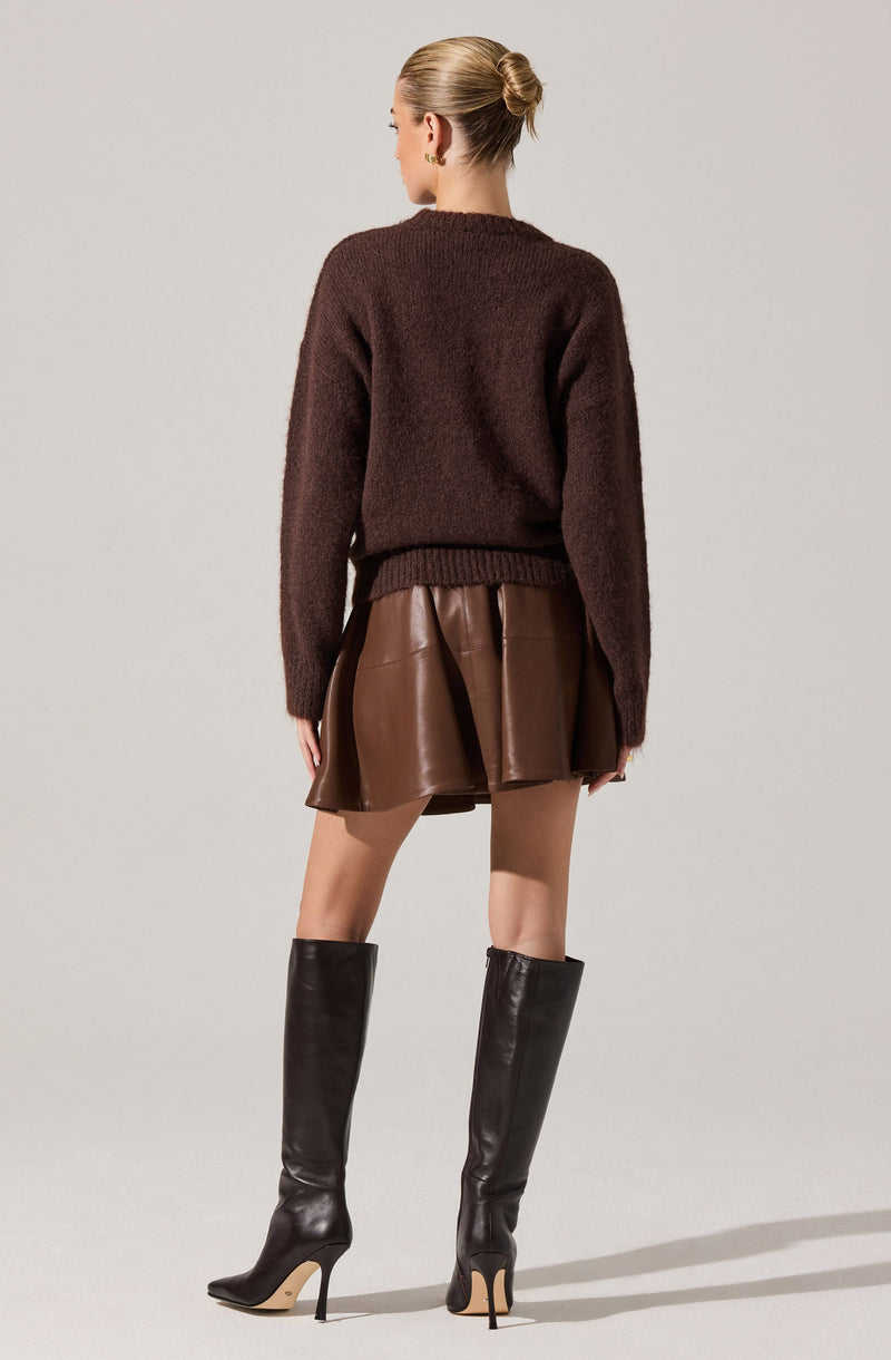 A woman is wearing a loose, dark brown knit sweater with a ribbed hem and cuffs. She pairs it with a shiny, brown leather mini skirt that has pleats. She completes her outfit with tall, black leather high-heeled boots.