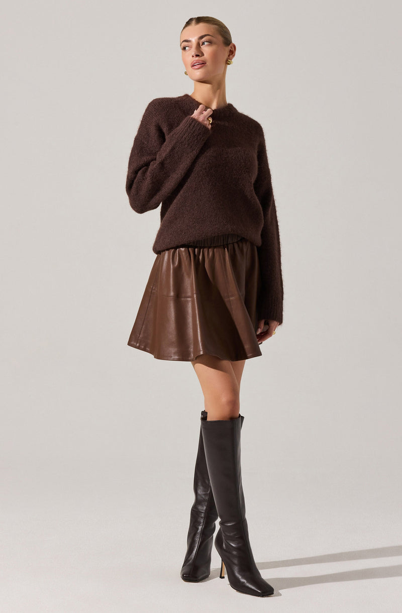 A woman is wearing a dark brown, fuzzy knit sweater with long sleeves and a rounded neckline. She pairs it with a high-waisted, brown leather skirt that has a flared silhouette. She completes the look with tall, black leather heeled boots that reach just below the knee.