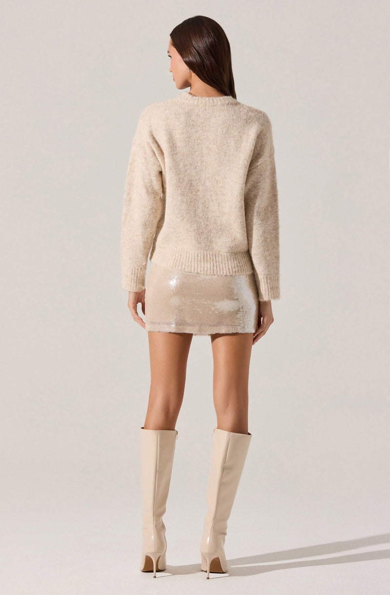 The woman is wearing a loose, long-sleeved, beige knitted sweater with a round neckline. She has on a short, shiny, beige sequin skirt and tall, matching beige thigh-high boots with high heels.