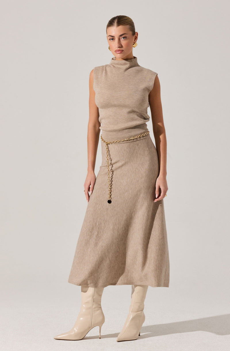 The woman is wearing a sleeveless, high-neck beige top made of a soft, knit fabric, paired with a matching long beige skirt that reaches above her ankles. She accessorizes with a gold chain belt around her waist and gold hoop earrings. She completes her outfit with off-white, shiny, knee-high heeled boots.