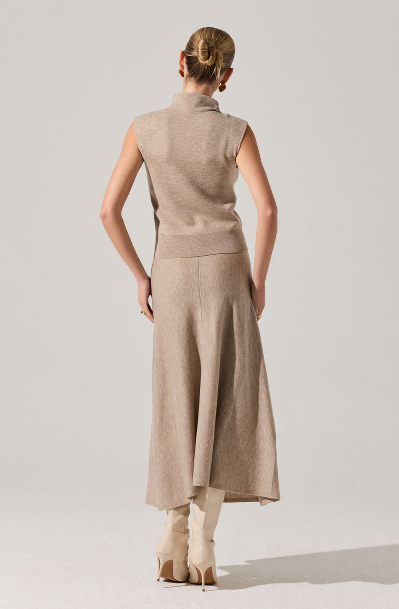 A woman is wearing a sleeveless, high-neck beige vest and a matching long, wide-leg skirt made of a soft, textured fabric. She has her hair styled in a low bun and is standing with her back facing the camera. She is also wearing knee-high beige boots with high heels.