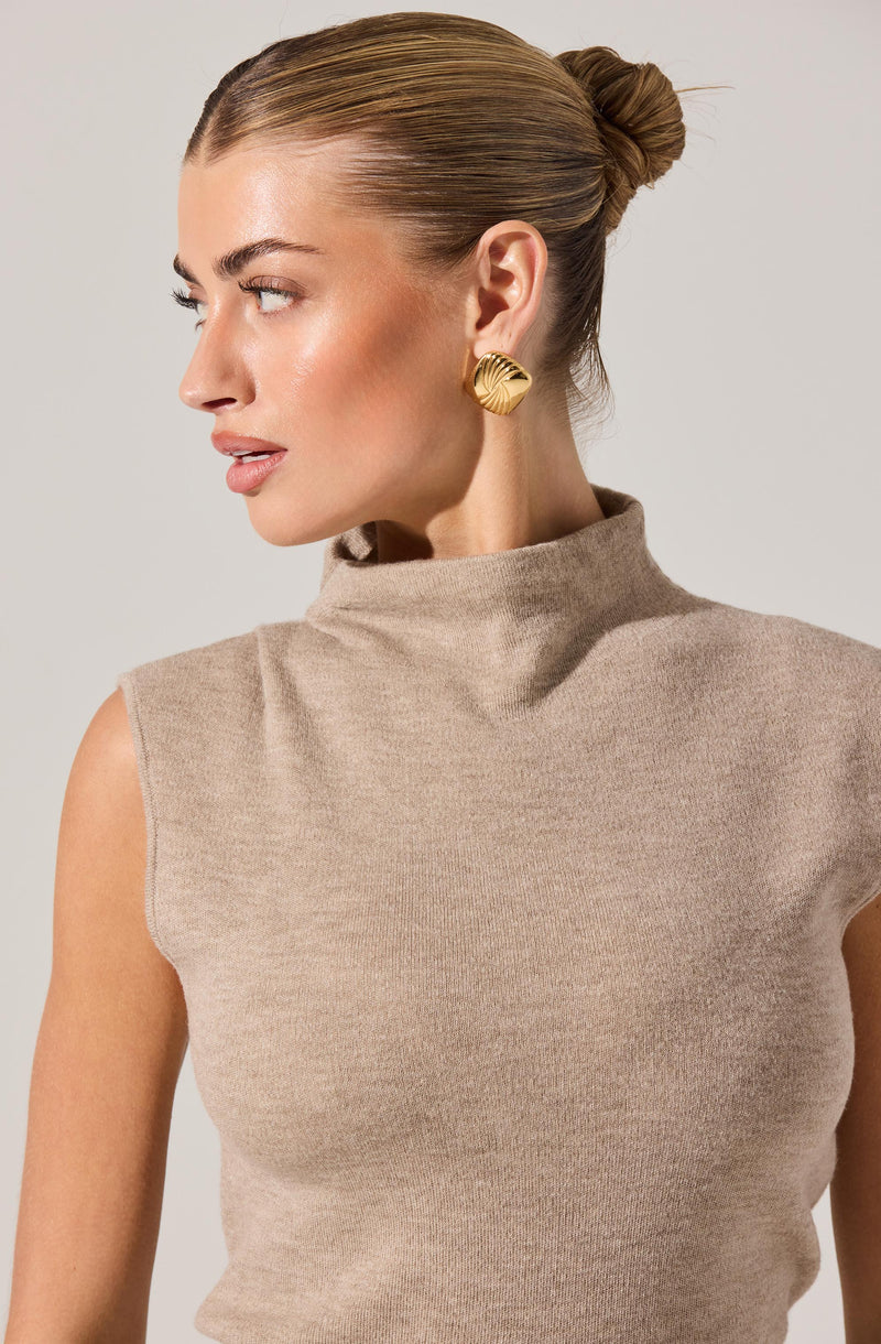 A woman is wearing a sleeveless, high-neck beige knit top. She has a large, gold, shell-shaped earring on her visible ear and her hair is styled in a sleek, tight bun.