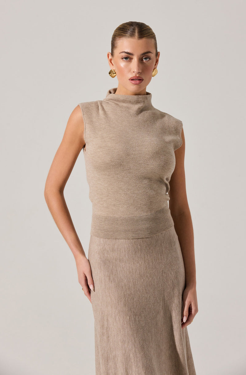 A woman is wearing a sleeveless beige knit top with a high neckline and a matching beige knit skirt that reaches below the knees. The top is fitted at the waist with a ribbed band, and the skirt has a subtle ribbed texture. She accessorizes with gold earrings.