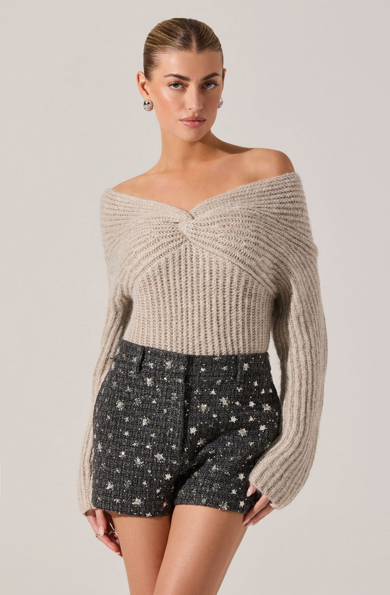 A woman is wearing a beige, off-the-shoulder knit sweater with a twisted front detail and long sleeves. She is also wearing high-waisted, black textured shorts with a white floral pattern.