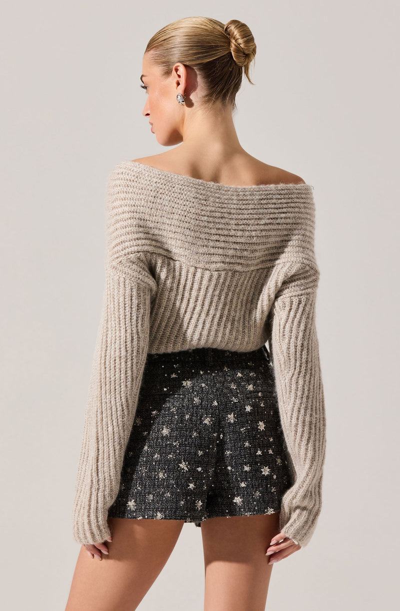 A woman is wearing a beige, chunky knit sweater with wide horizontal ribbing on the upper part and vertical ribbing on the sleeves. She pairs it with a black, textured skirt that features a pattern of small, shiny, star-like embellishments.