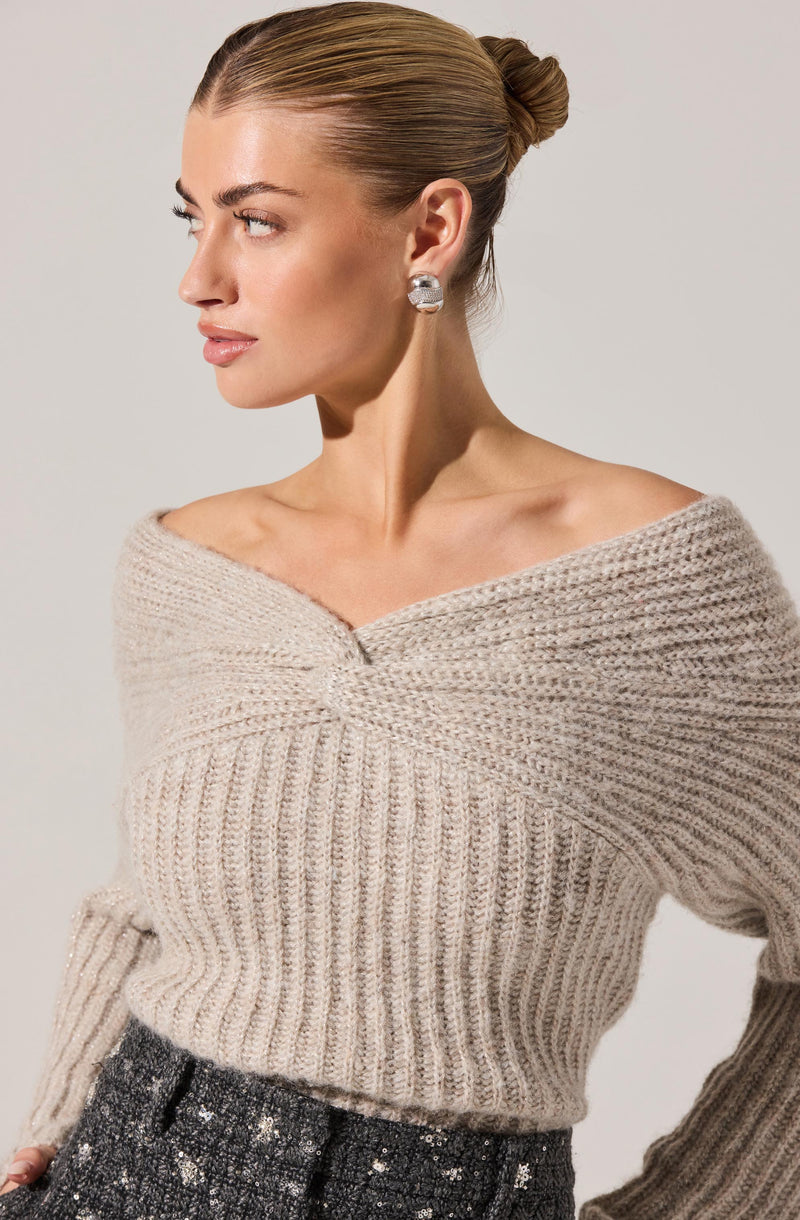 A woman is wearing a beige, chunky knit sweater with a wide neckline that drapes off her shoulders. The sweater has a ribbed pattern throughout. She is also wearing sparkling, large, circular earrings and a dark, textured skirt with metallic accents.