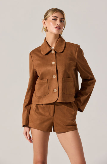 A woman is wearing a matching brown suede jacket and shorts set. The jacket has a rounded collar, four large buttons down the front, and two front pockets. The shorts are high-waisted and have a tailored fit.