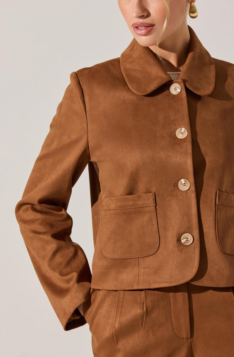 A woman is wearing a brown, tailored jacket made of a faux suede material. The jacket features a rounded collar, four large buttons down the front, and two patch pockets on each side at the front. The sleeves are long, and she has one hand in her pocket.