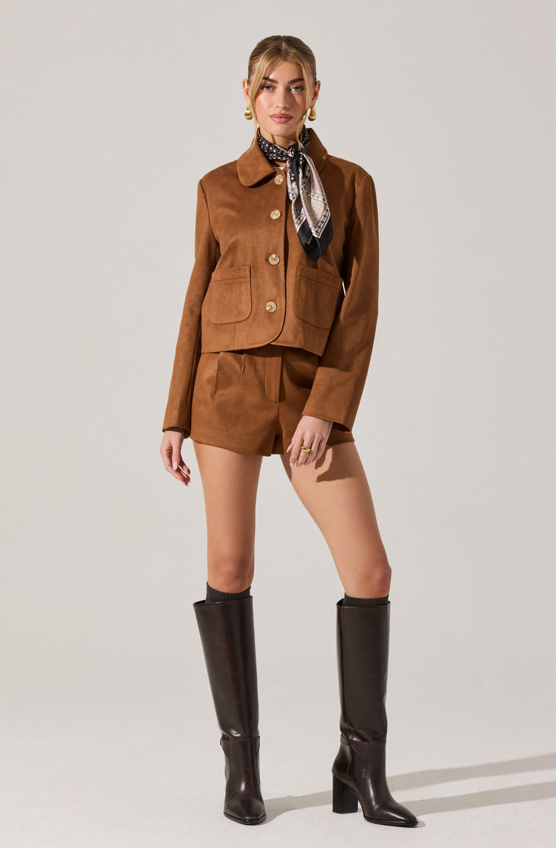 A woman is wearing a brown suede jacket with large buttons and patch pockets, paired with matching high-waisted shorts. She accessorizes with a black and white patterned scarf tied around her neck, large gold hoop earrings, and gold rings. She is also wearing tall black knee-high boots.
