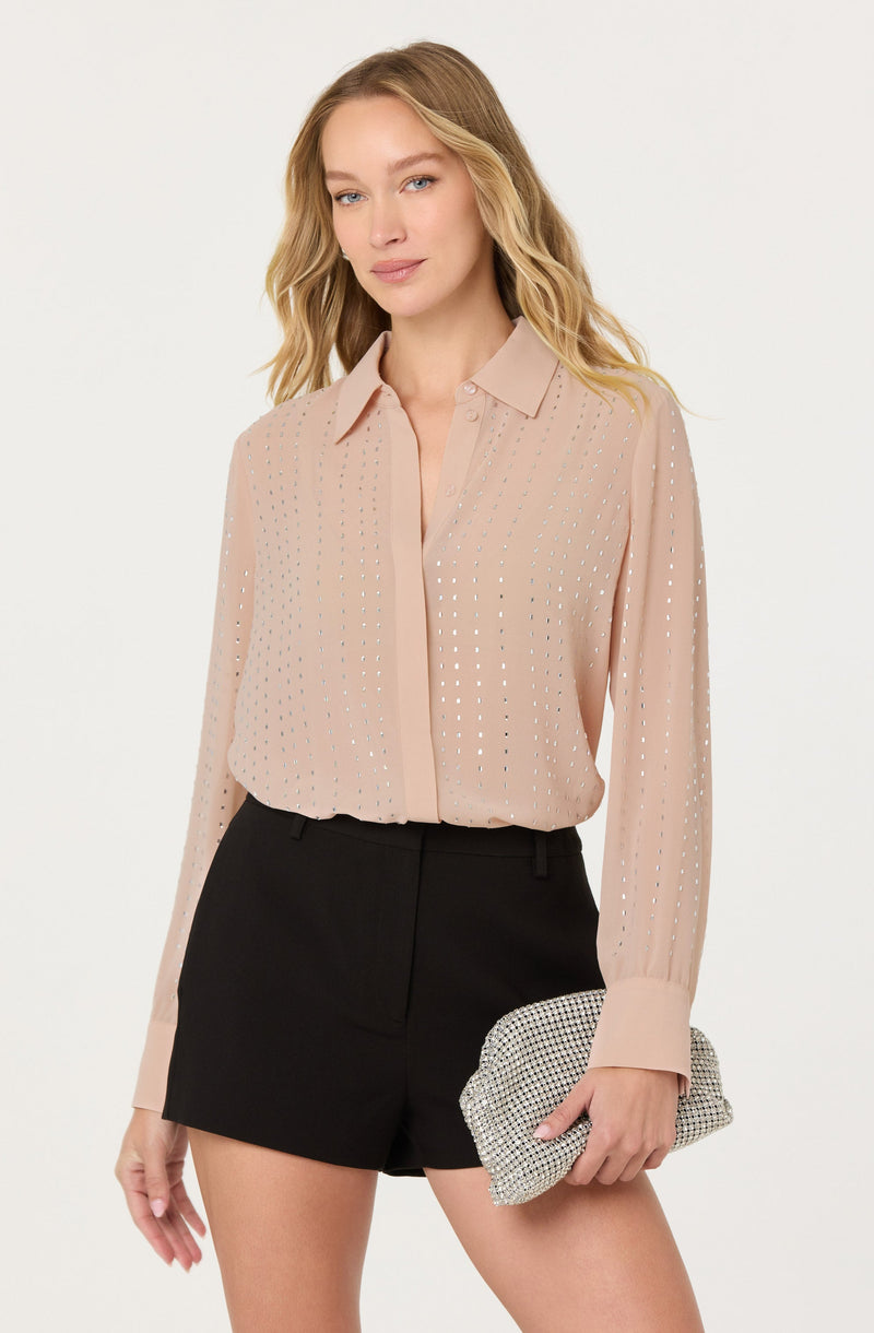 A woman is wearing a light pink, long-sleeved blouse with a collar and vertical perforated pattern throughout. The blouse has a loose fit with a hidden button placket down the front and wide cuffs at the sleeves. She pairs it with black high-waisted shorts. She is holding a small, rectangular, patterned clutch in her right hand.