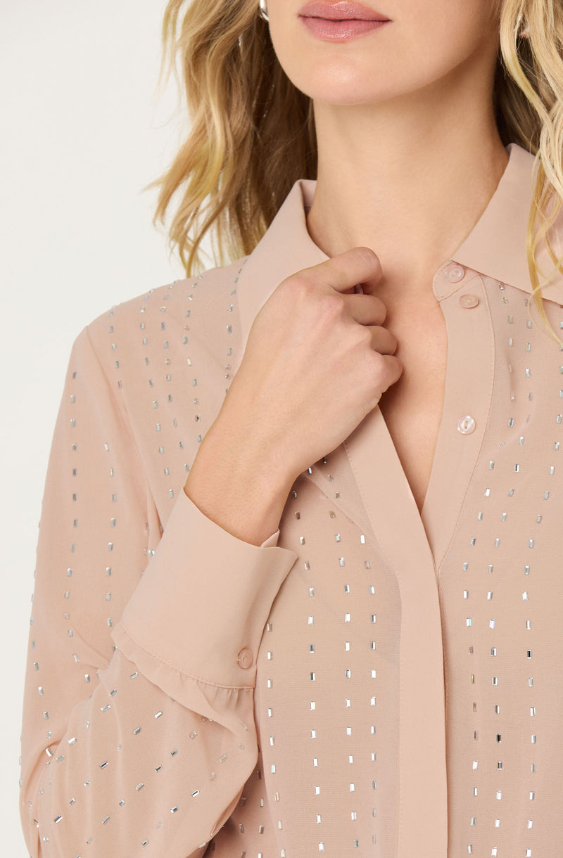 A woman is wearing a light beige, long-sleeved blouse with a pointed collar. The blouse features vertical rows of small, shiny, silver rectangular embellishments across the fabric. The sleeves are buttoned at the cuffs, and the top buttons of the blouse are undone, revealing a small portion of the neckline.