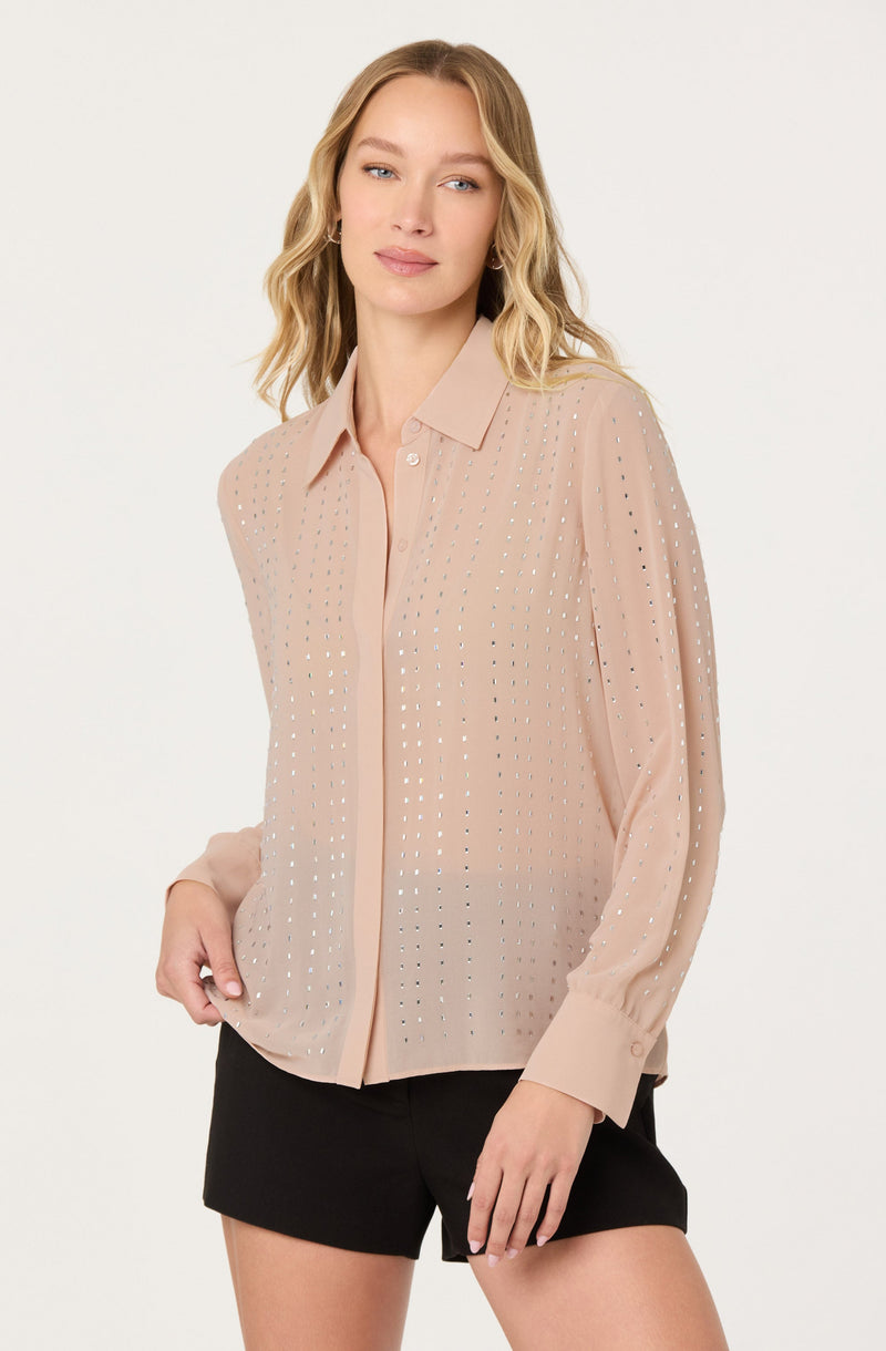 A woman is wearing a light beige long-sleeve button-up shirt with a collar. The shirt features a pattern of small, evenly spaced metallic or shiny dots throughout. She has paired the shirt with black shorts.