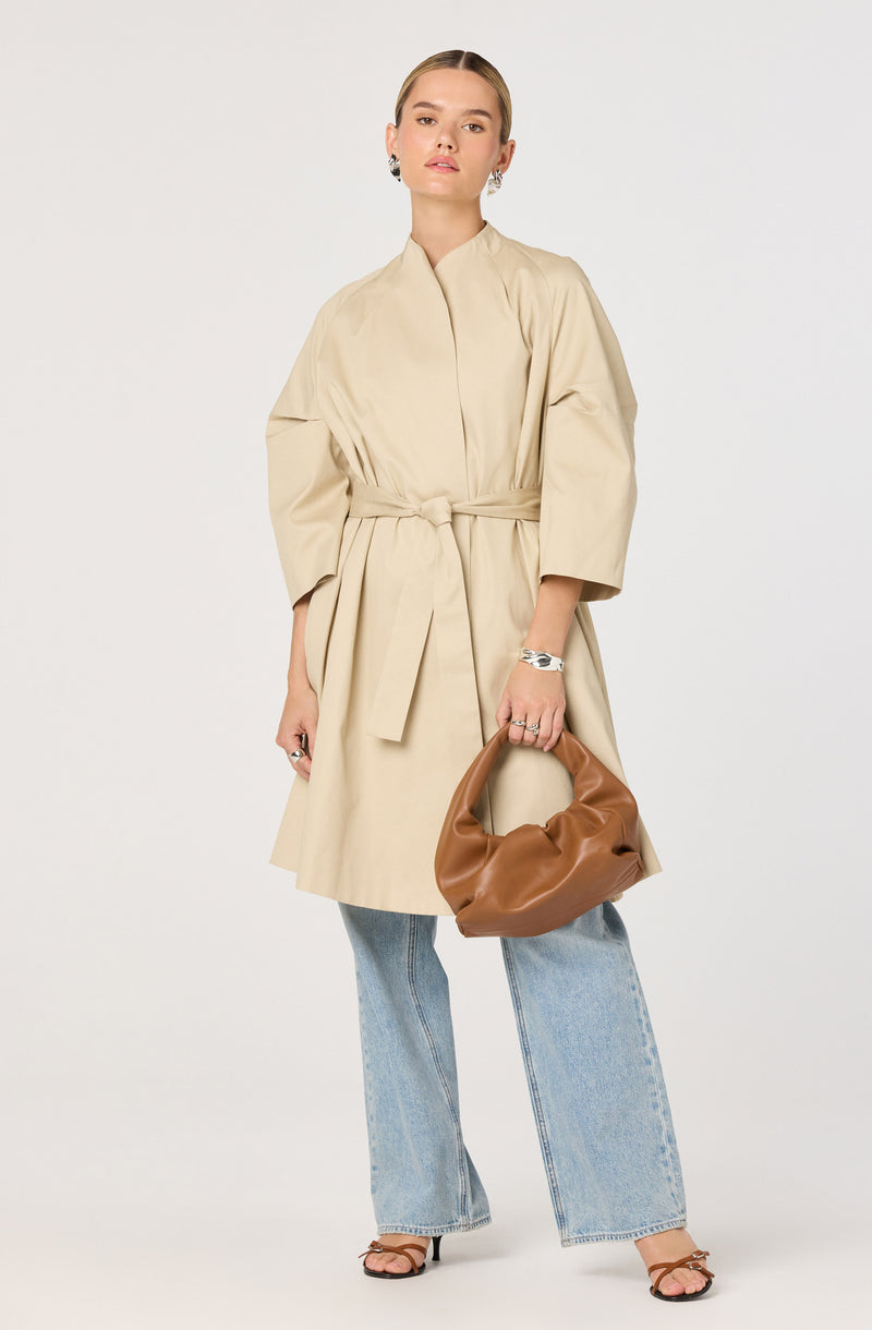 A woman wears a beige, knee-length coat with wide sleeves and a waist tie. She pairs the coat with loose-fitting, light-wash jeans and strappy high-heeled sandals. She accessorizes with earrings, a bracelet, and rings, and holds a brown, soft, slouchy handbag.