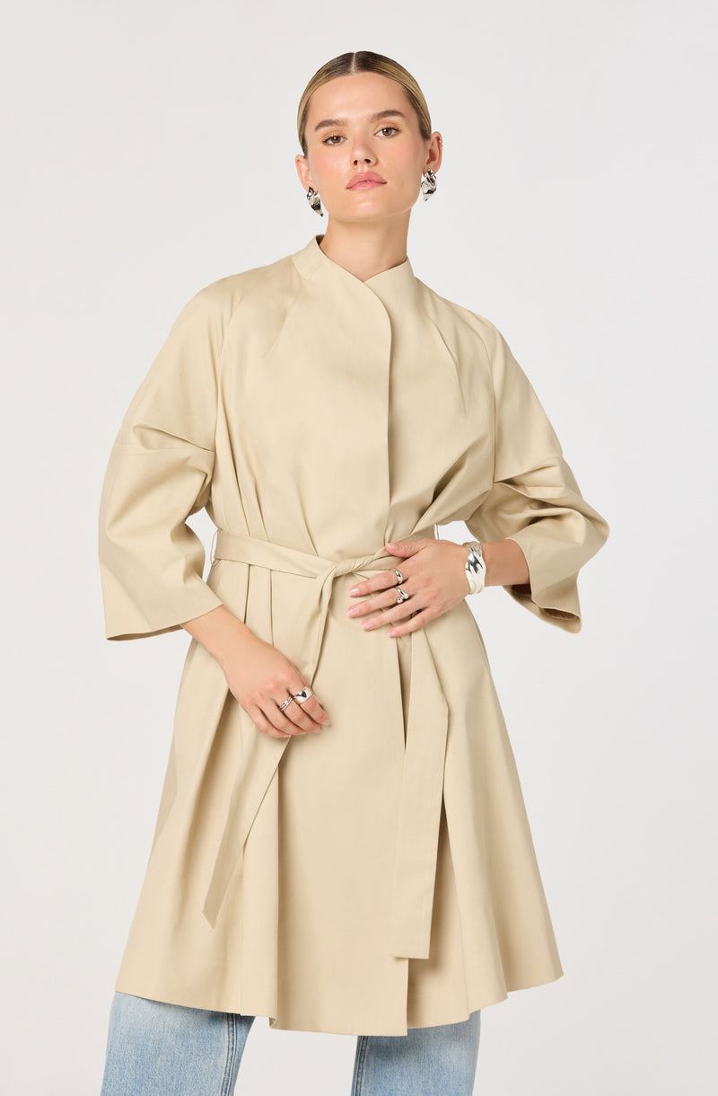 A woman wearing a beige, knee-length coat with a matching fabric belt tied at the waist. The coat has a high collar and three-quarter length puffed sleeves. She is accessorized with large earrings, rings, and a bracelet.