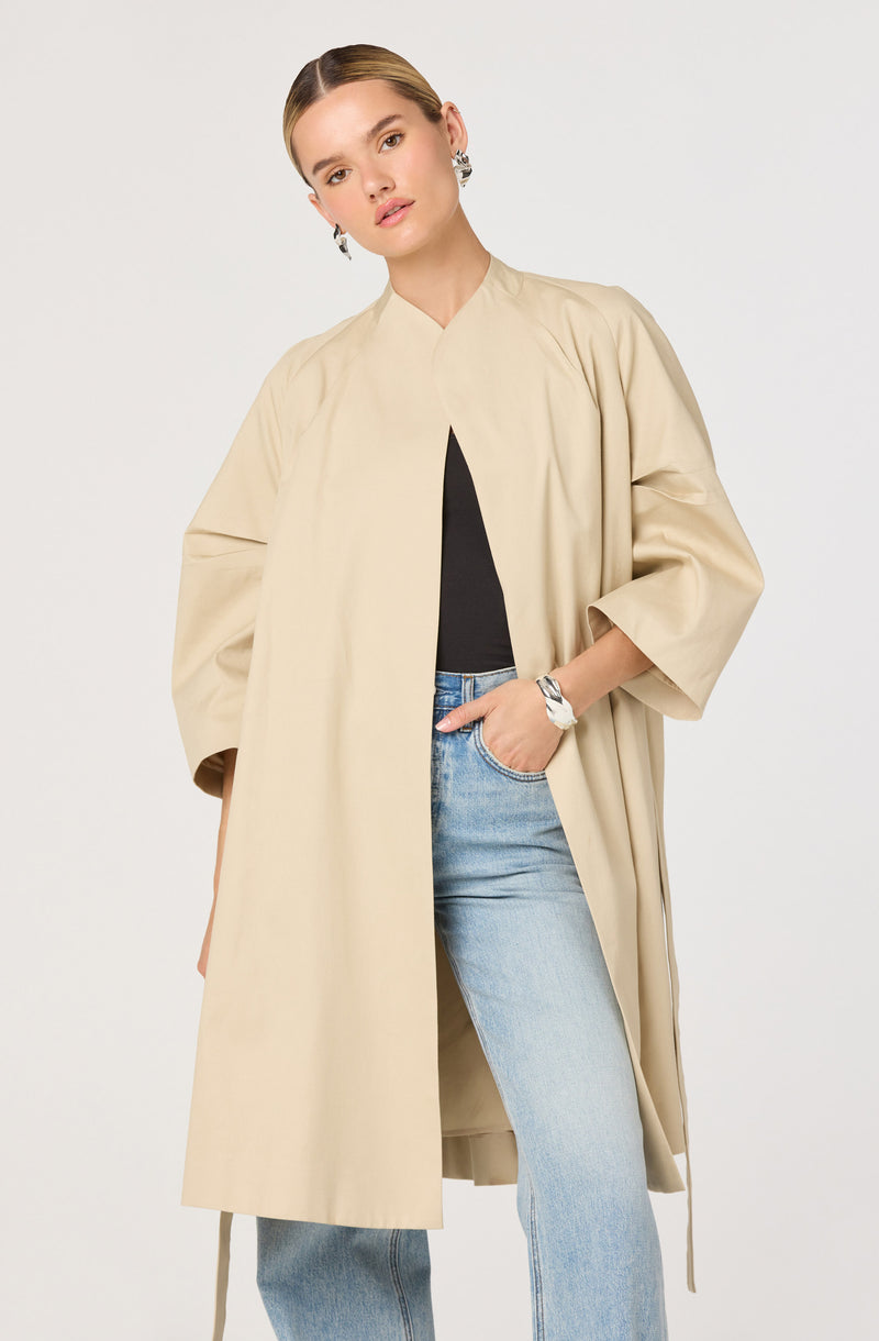 A woman is wearing a knee-length, loose-fitting beige coat with wide three-quarter sleeves. Underneath, she has on a black top, and she is also wearing light blue high-waisted jeans. She has her right hand in her pocket and is accessorized with large, round earrings and a bracelet on her left wrist.