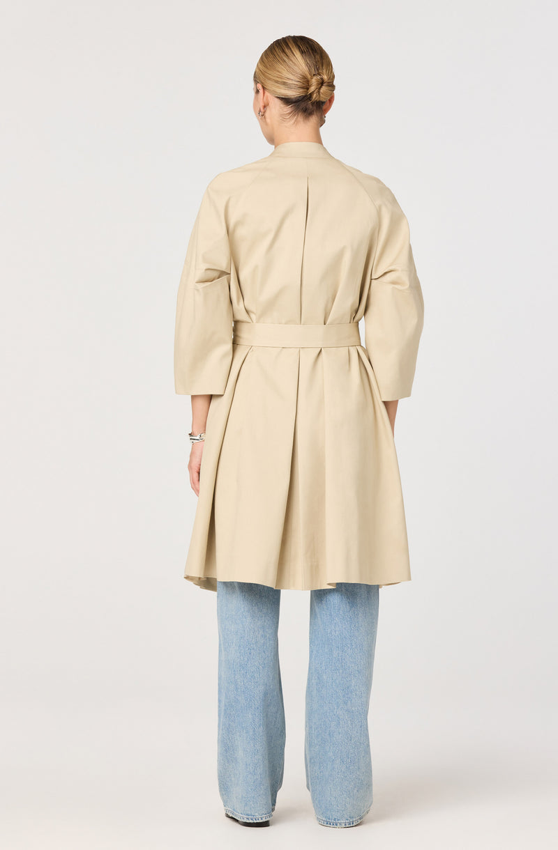 A person is wearing a beige, knee-length trench coat with wide three-quarter sleeves, a fitted waist, and a pleated skirt. The coat has a high collar and appears to be made of a lightweight fabric. The person is also wearing light blue, straight-leg jeans. Their hair is styled in a low bun.