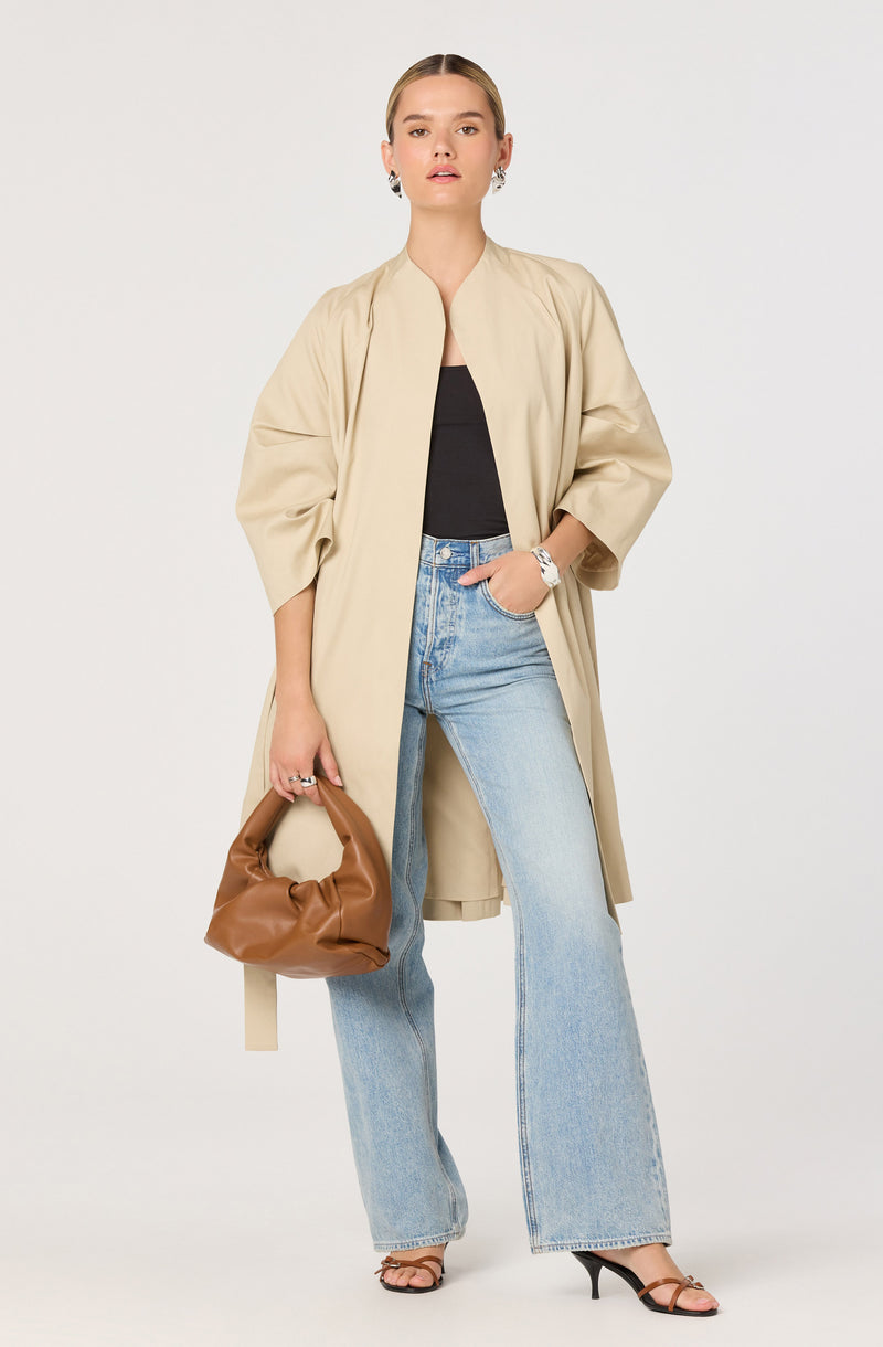 A woman is wearing a long beige coat with wide sleeves over a black top. She has on light-wash high-waisted jeans with a relaxed fit. She accessorizes with earrings, a watch, and a ring. She is holding a brown, soft, gathered handbag and is wearing black strappy high-heeled sandals.