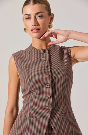 A woman is wearing a sleeveless, fitted dress with a high neckline and a small checkered pattern in shades of brown and black. The dress features a row of fabric-covered buttons down the front and two small pockets on the lower front. She is accessorized with large, gold, geometric earrings.