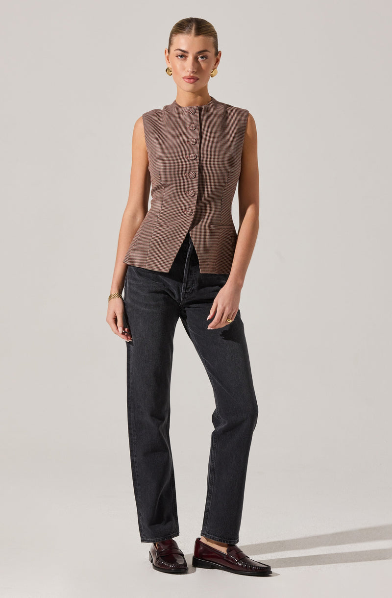 A woman is wearing a sleeveless, button-up vest with a small, checkered pattern in shades of brown and beige. She pairs it with high-waisted, dark-wash jeans and dark brown loafers. She accessorizes with gold jewelry, including earrings, a bracelet, and rings.