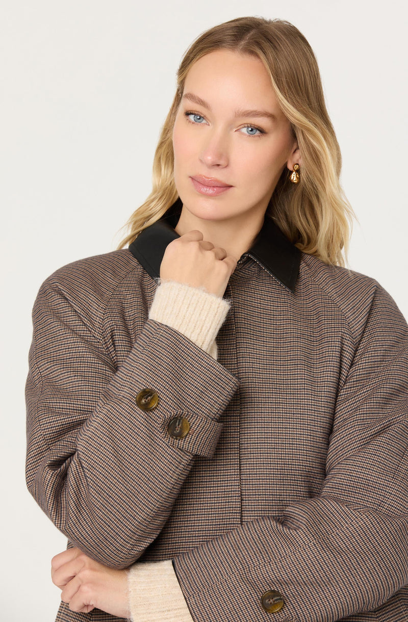 A woman wearing a brown plaid blazer with dark buttons, a black collar, and cream-colored knit cuffs. She has blonde, wavy hair and is accessorized with gold hoop earrings.