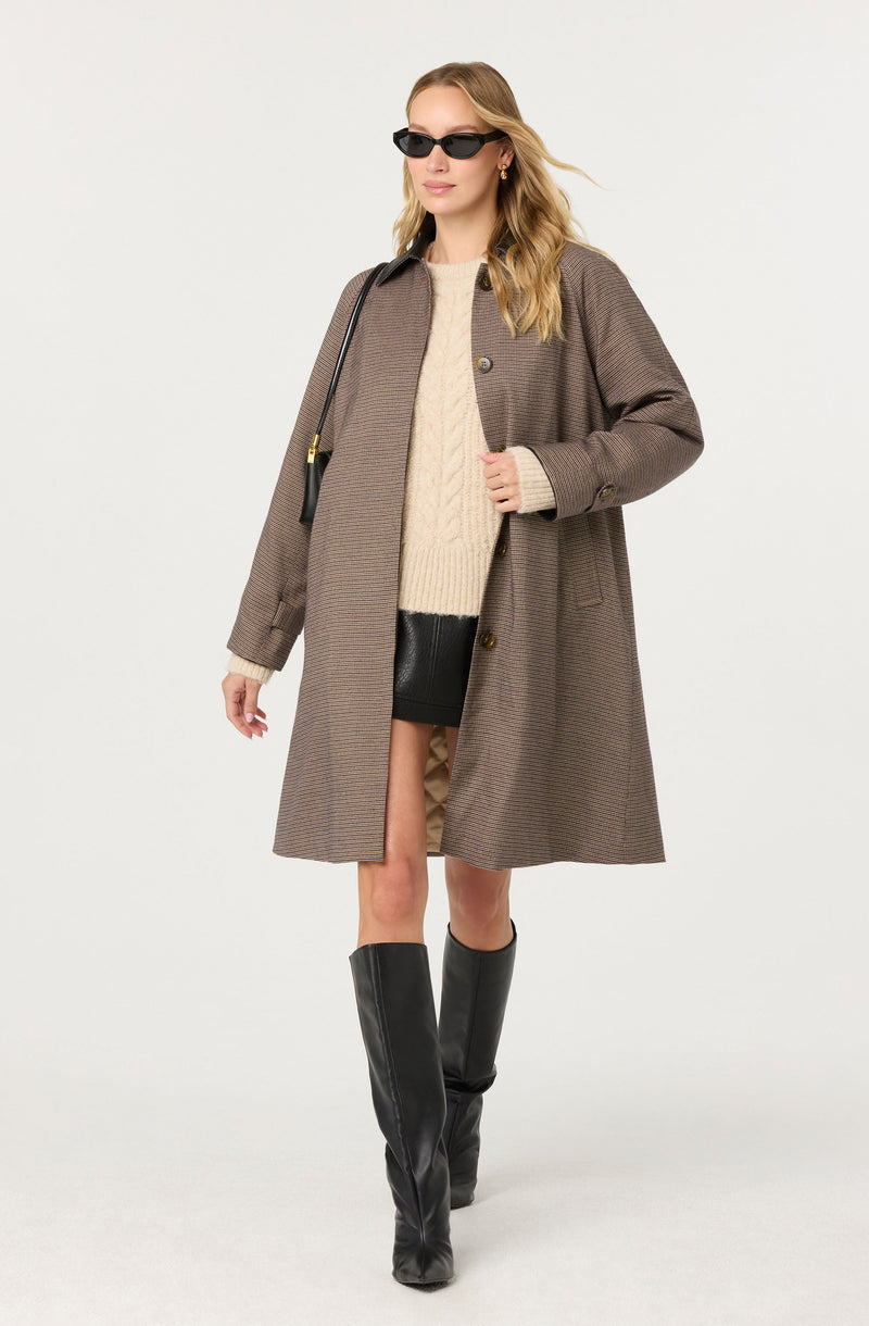 A woman is wearing a long brown checkered coat with a collar and large buttons. Underneath, she has on a cream-colored cable-knit sweater. She is also wearing black leather shorts that are partly visible below the coat. She has on black knee-high boots with a slight heel. She accessorizes with black sunglasses, small hoop earrings, and a black shoulder bag with a gold chain strap.