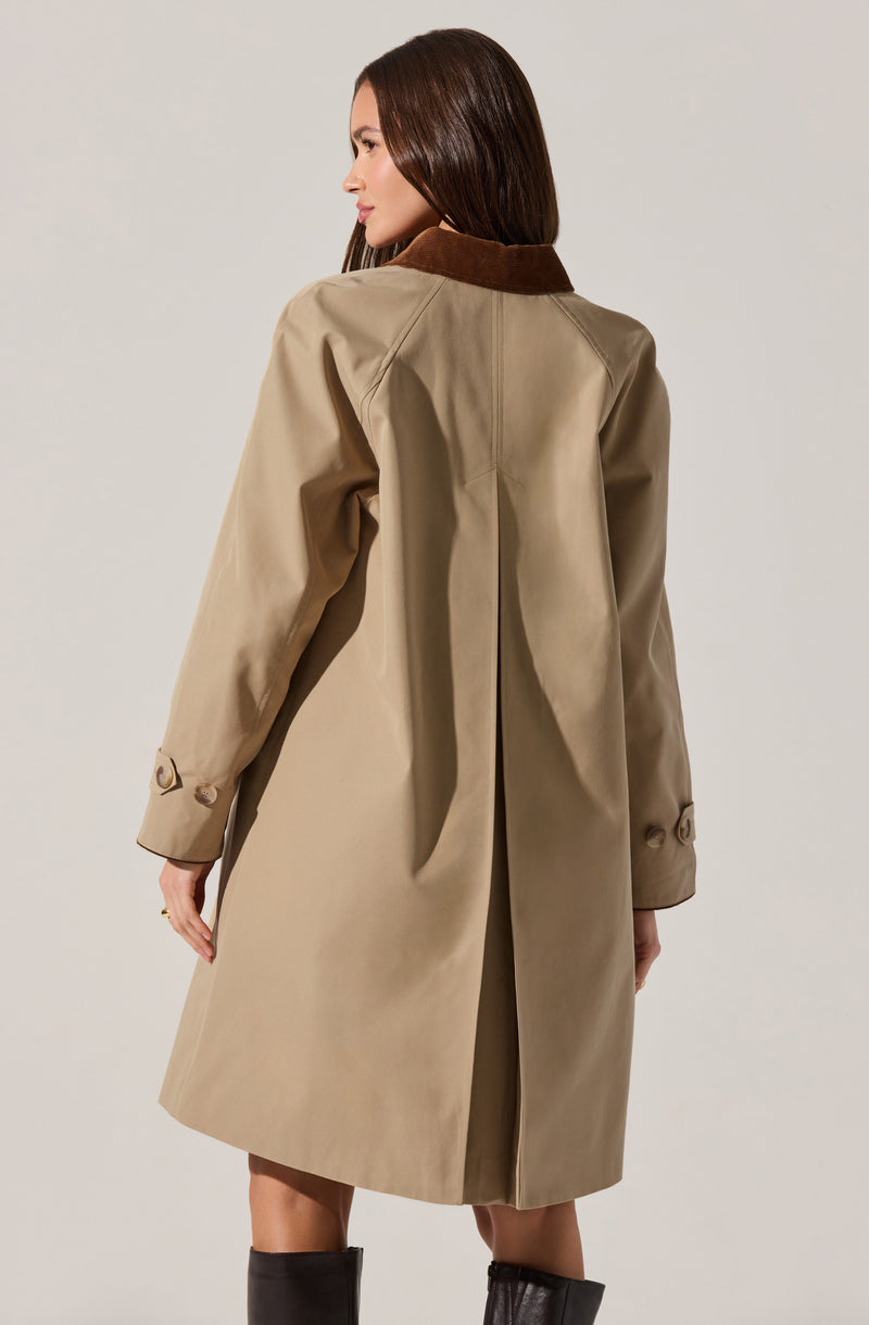 A woman is wearing a beige trench coat with a brown collar. The coat has long sleeves with decorative buttons near the cuffs, and it features a pleat at the back for movement. The coat is knee-length and appears to be made of a classic, smooth fabric.