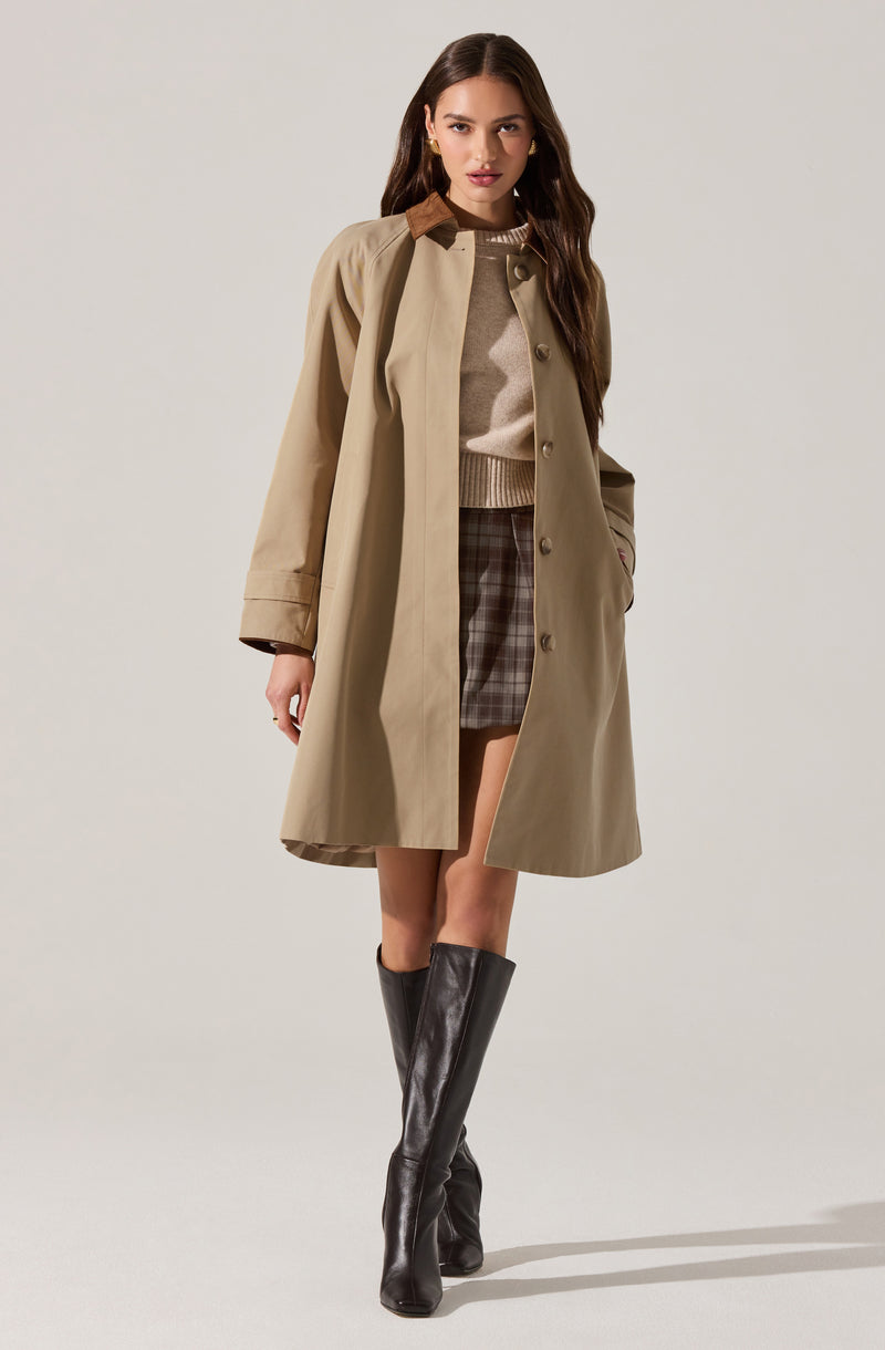 A woman is wearing a beige trench coat with a brown collar, buttoned down the front. Underneath, she has on a light beige sweater with a ribbed hem. She pairs this with a plaid skirt in shades of brown and gray. She is also wearing knee-high black leather boots.