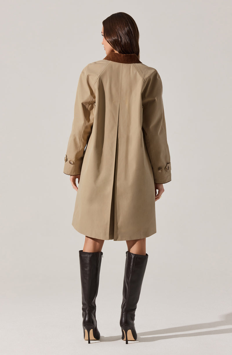 A woman is wearing a beige trench coat with a brown collar. The coat has long sleeves with buttoned straps near the cuffs, and it extends down to her knees. She is also wearing tall, black high-heeled leather boots.