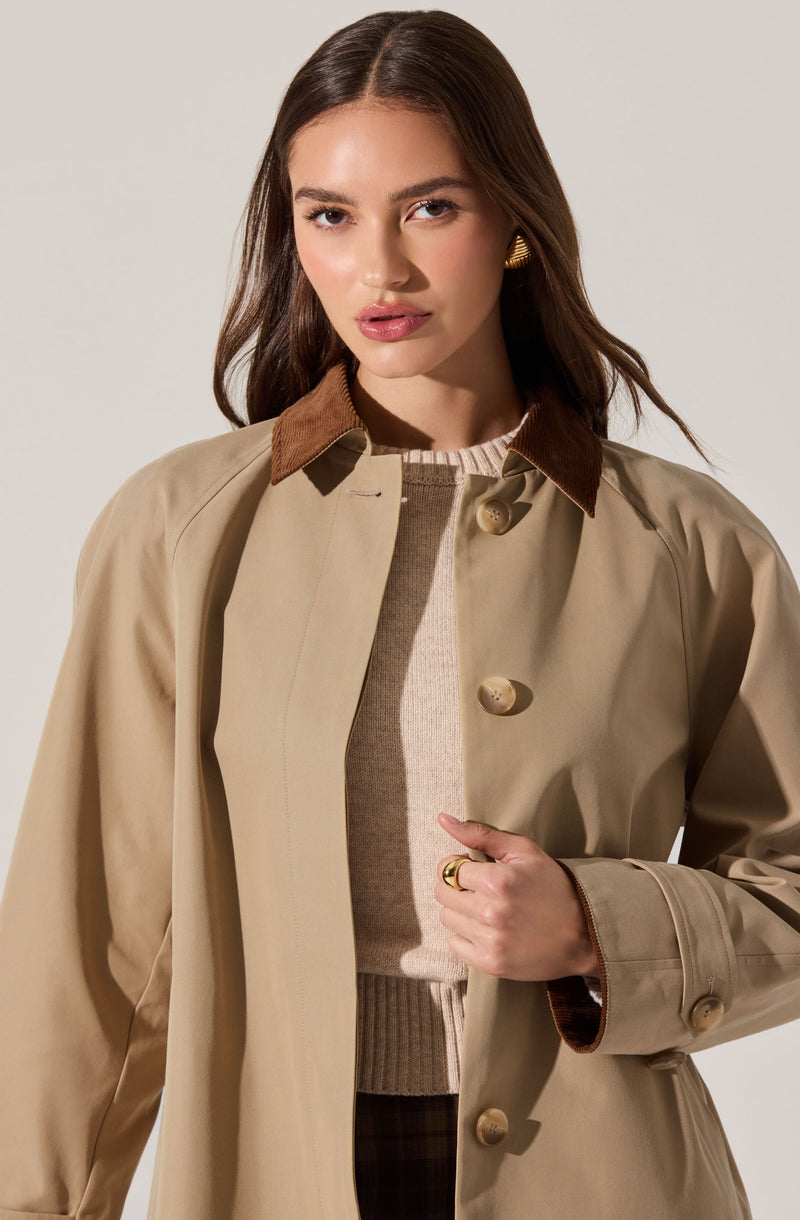 A woman is wearing a beige trench coat with a brown corduroy collar, beige buttons down the front, and a belt at the waist. Underneath, she has on a light-colored sweater.