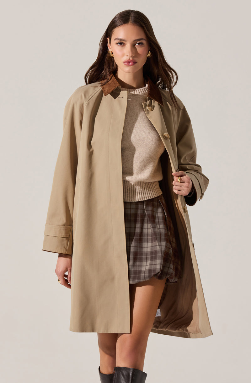 A woman is wearing a beige trench coat with a brown corduroy collar, a light beige turtleneck sweater, and plaid shorts in shades of brown and gray. She has gold earrings and rings on her fingers, and is also wearing black knee-high boots.