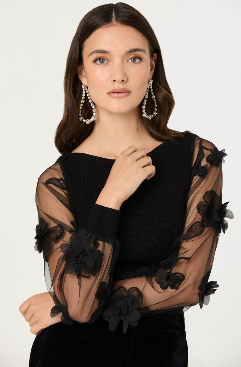 A woman is wearing a black top with sheer, long sleeves that feature black 3D floral appliqués. The main body of the top is made of a ribbed fabric, while the sleeves are transparent with embroidered flowers. She accessorizes with large, dangling earrings adorned with clear, sparkling stones.
