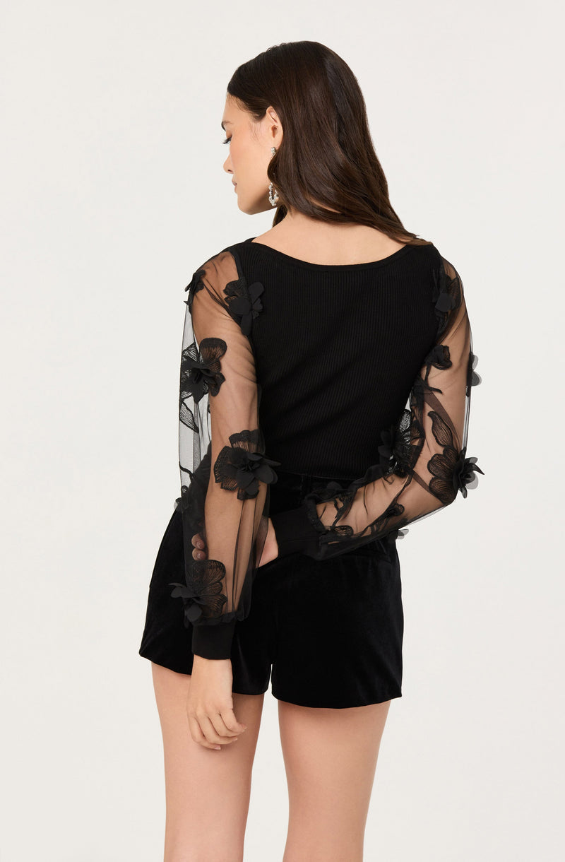 A woman is wearing a black long-sleeve top with sheer, transparent sleeves adorned with 3D floral embroidery, paired with black shorts. The top has a fitted, ribbed bodice, and the flowers on the sleeves are made of embroidered fabric, giving a textured, decorative look.
