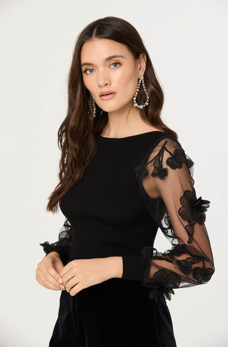 A woman is wearing a black top with sheer long sleeves that feature floral embroidery and three-dimensional flower appliqués. The top has a round neckline and fitted body. She is also accessorized with large, chandelier-style earrings.