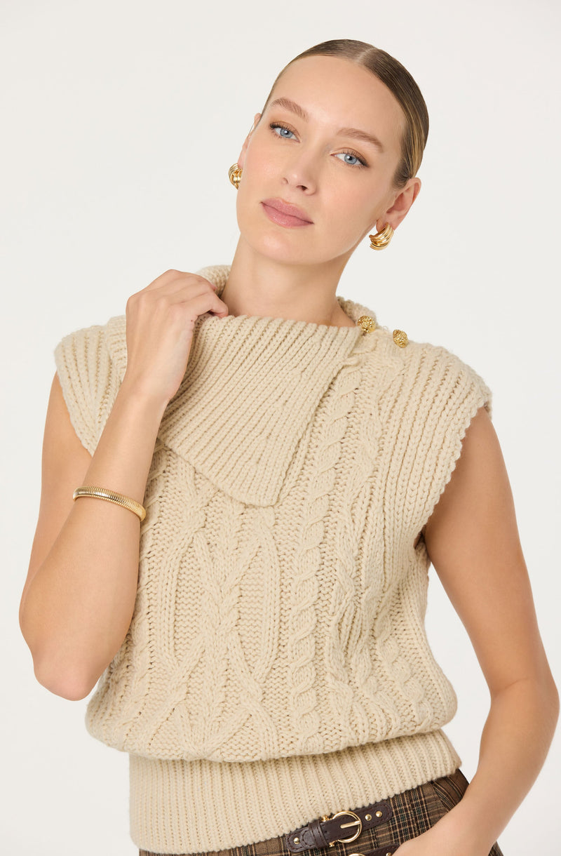 A woman is wearing a sleeveless, chunky knit sweater in a light cream color with a large, folded collar. The sweater features a cable knit pattern and has a ribbed waistband. She accessorizes with gold hoop earrings, a gold bracelet, and a gold pin on the collar, and she has her hair slicked back.