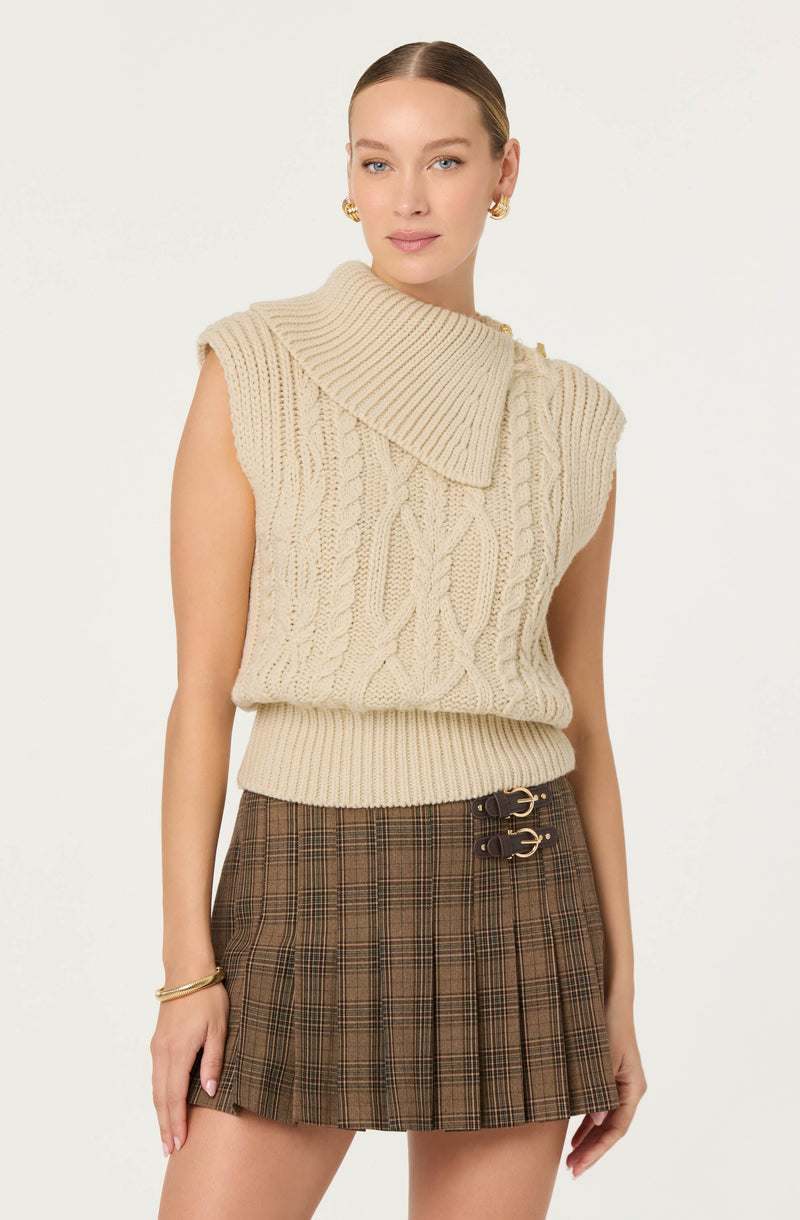A woman is wearing a sleeveless, chunky knit, cream-colored sweater with a large, ribbed, asymmetrical collar. The sweater has a textured cable knit pattern. She pairs it with a brown plaid skirt featuring a high waist and decorative buckles on the front. She accessorizes with gold hoop earrings and a gold bracelet on her left wrist.