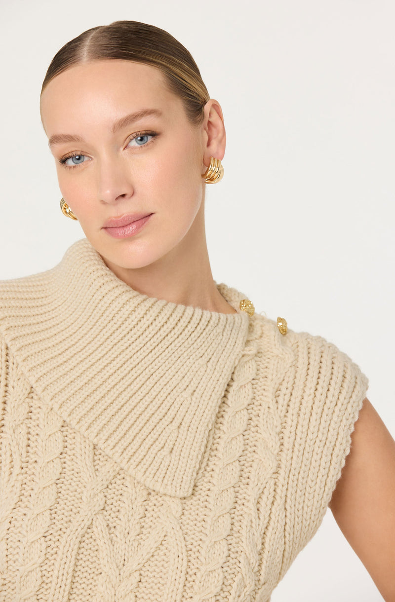 A woman with fair skin and blonde hair styled in a sleek, low bun is wearing large gold hoop earrings and a chunky, cream-colored, cable-knit sleeveless sweater with a high, fold-over collar and decorative gold buttons on the shoulder.
