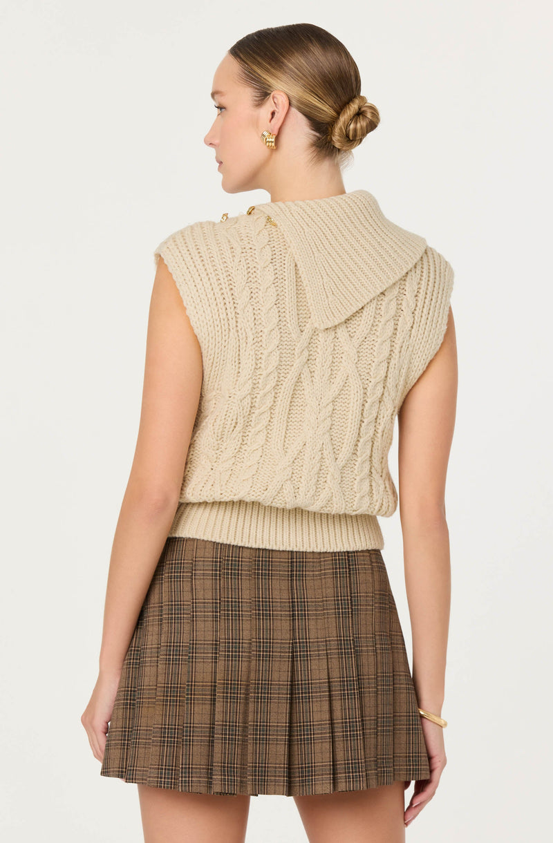 A woman is wearing a sleeveless, chunky beige knit sweater with a large collar that drapes over her shoulders. The sweater has a cable knit pattern and a ribbed hem. She pairs it with a brown plaid pleated mini skirt. She also has gold earrings and a gold bracelet, and her hair is styled in a low bun.