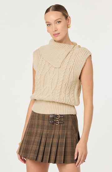 A woman is wearing a sleeveless cream-colored knit sweater with a high, rolled collar and cable-knit patterns. The sweater has decorative buttons on the shoulder. She pairs it with a brown plaid pleated skirt that features two decorative buckle straps at the waist.