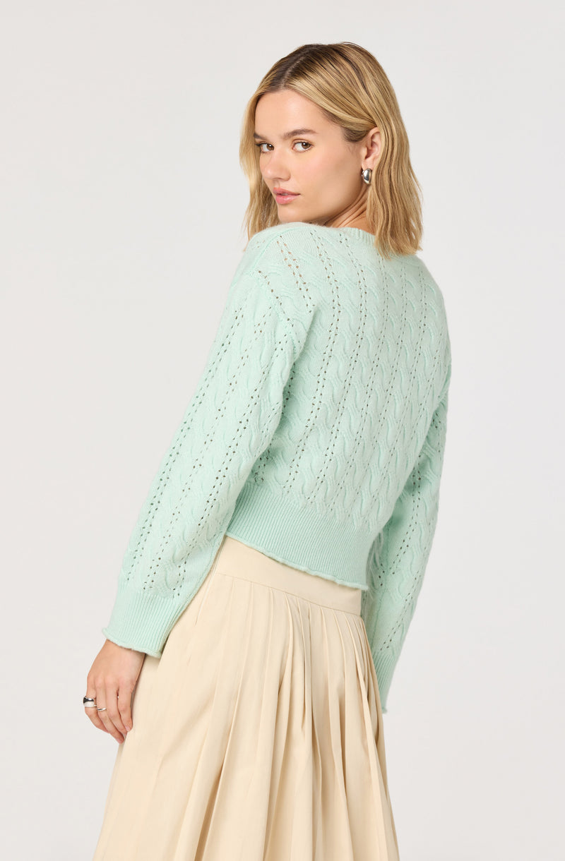 A woman is wearing a light mint green, long-sleeved knit sweater with an open, eyelet pattern. The sweater has a ribbed hem and cuffs, and the fabric appears soft and cozy. She pairs it with a high-waisted, cream-colored, pleated skirt that flows loosely. She also wears silver hoop earrings.