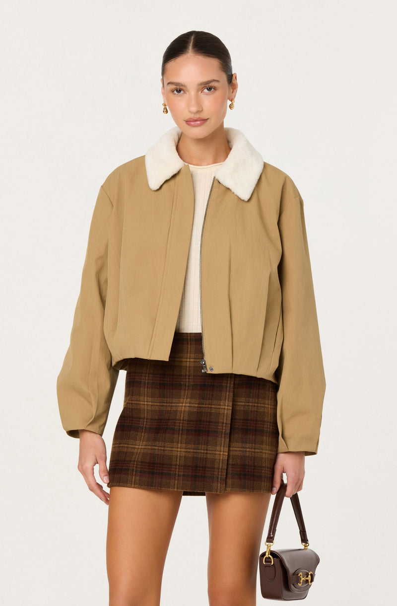 A woman is wearing a tan cropped jacket with a white faux fur collar. Underneath, she has on a light-colored knit sweater. She pairs these with a high-waisted, brown plaid mini skirt. She accessorizes with gold hoop earrings and carries a small brown handbag with a gold buckle.