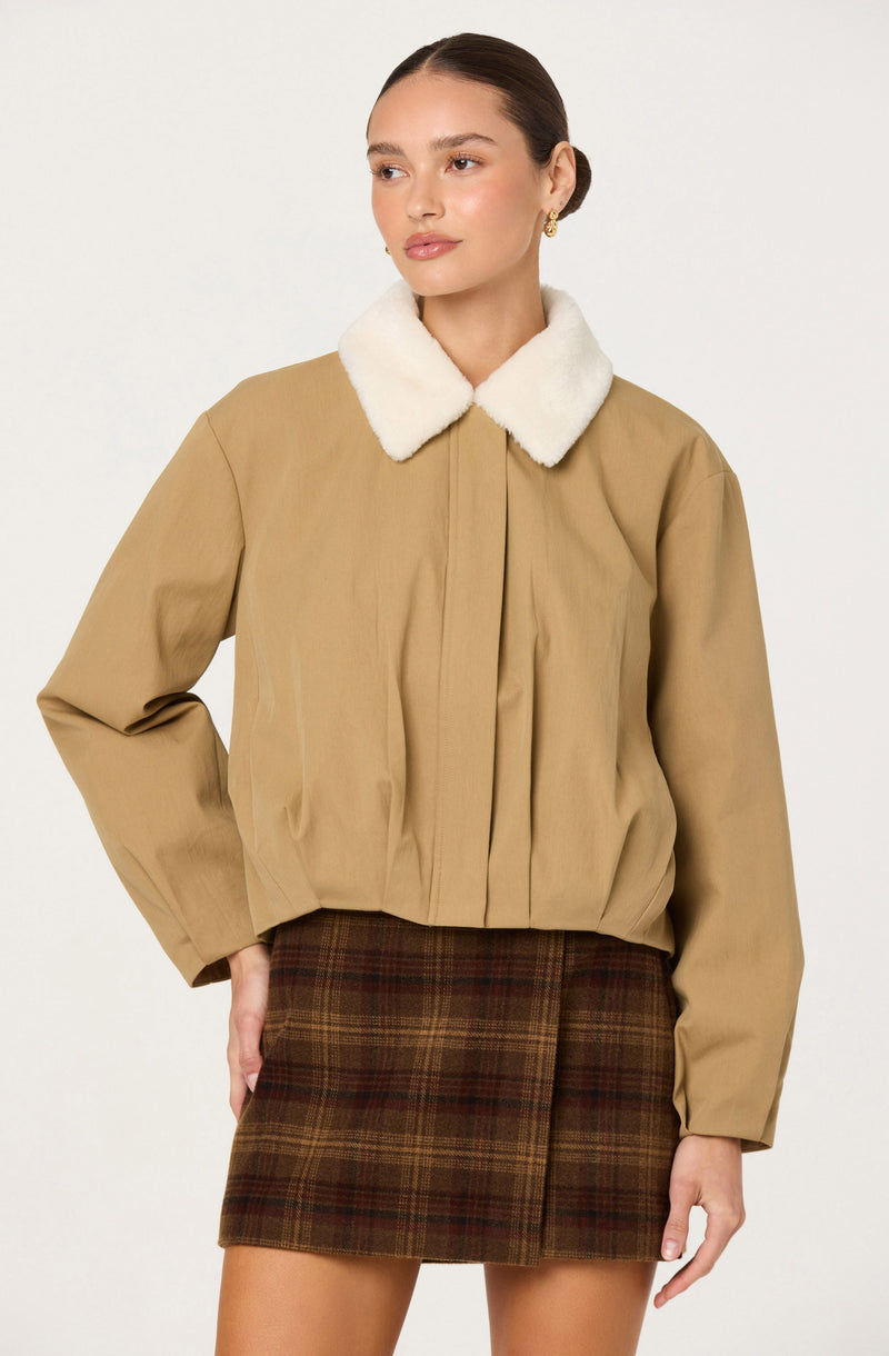 A woman is wearing a beige jacket with a white faux fur collar and long sleeves. She is also wearing a brown plaid skirt with a subtle pattern.