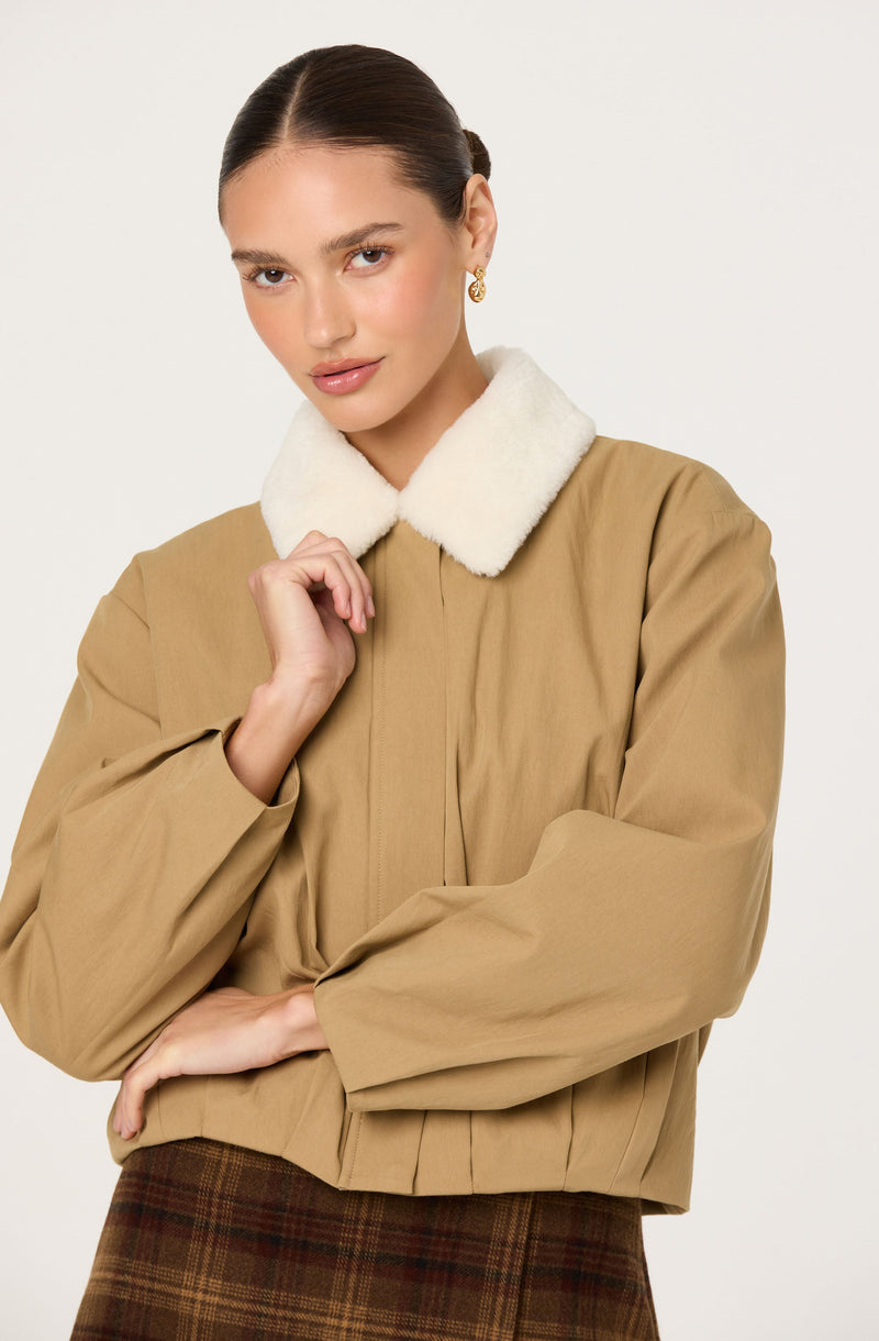 A woman is wearing a tan jacket with a white, fluffy collar. The jacket has a relaxed fit with gathered details at the waist and sleeves. She is also wearing earrings and a plaid skirt with brown and reddish tones.