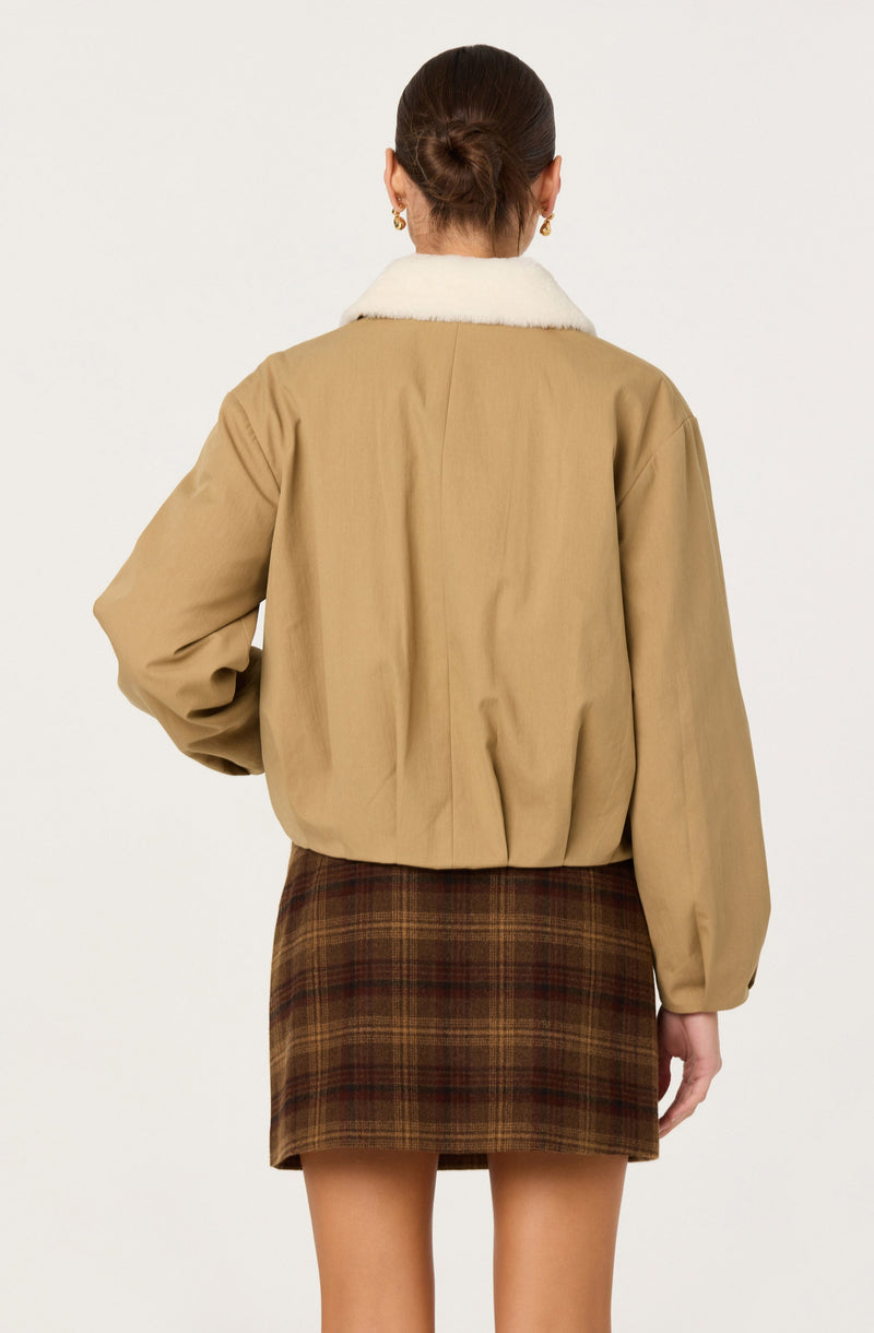 The image shows a woman wearing a beige jacket with a white, fluffy collar. The jacket has a slightly loose fit with gathered fabric at the cuffs. She is also wearing a short, brown plaid skirt with shades of darker brown and hints of red. Her hair is styled in a low bun, and she has small hoop earrings.