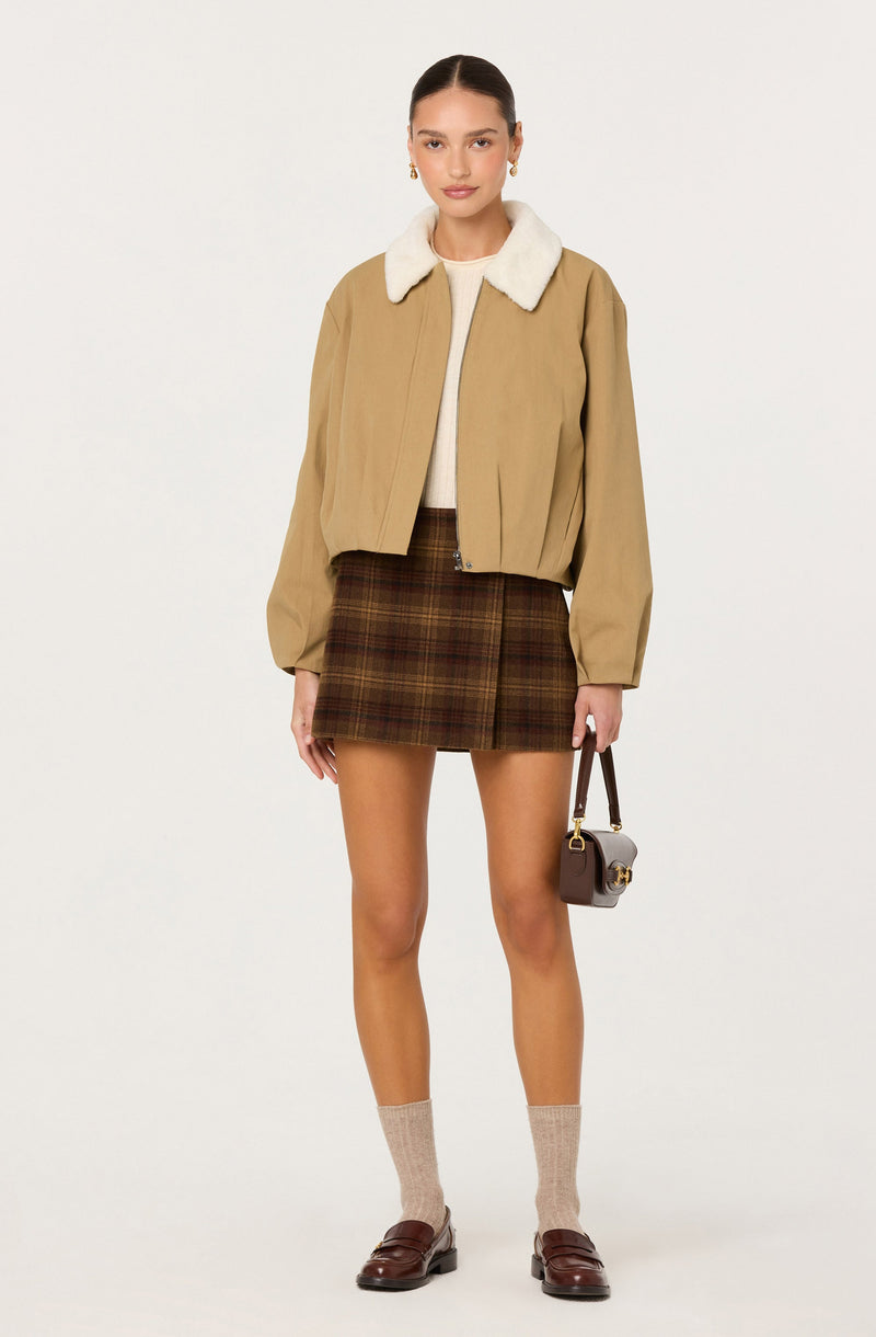 A woman is wearing a beige cropped jacket with a white faux fur collar, a cream-colored top underneath, a brown plaid skirt, beige socks, and brown loafers. She is holding a small brown handbag with gold hardware.